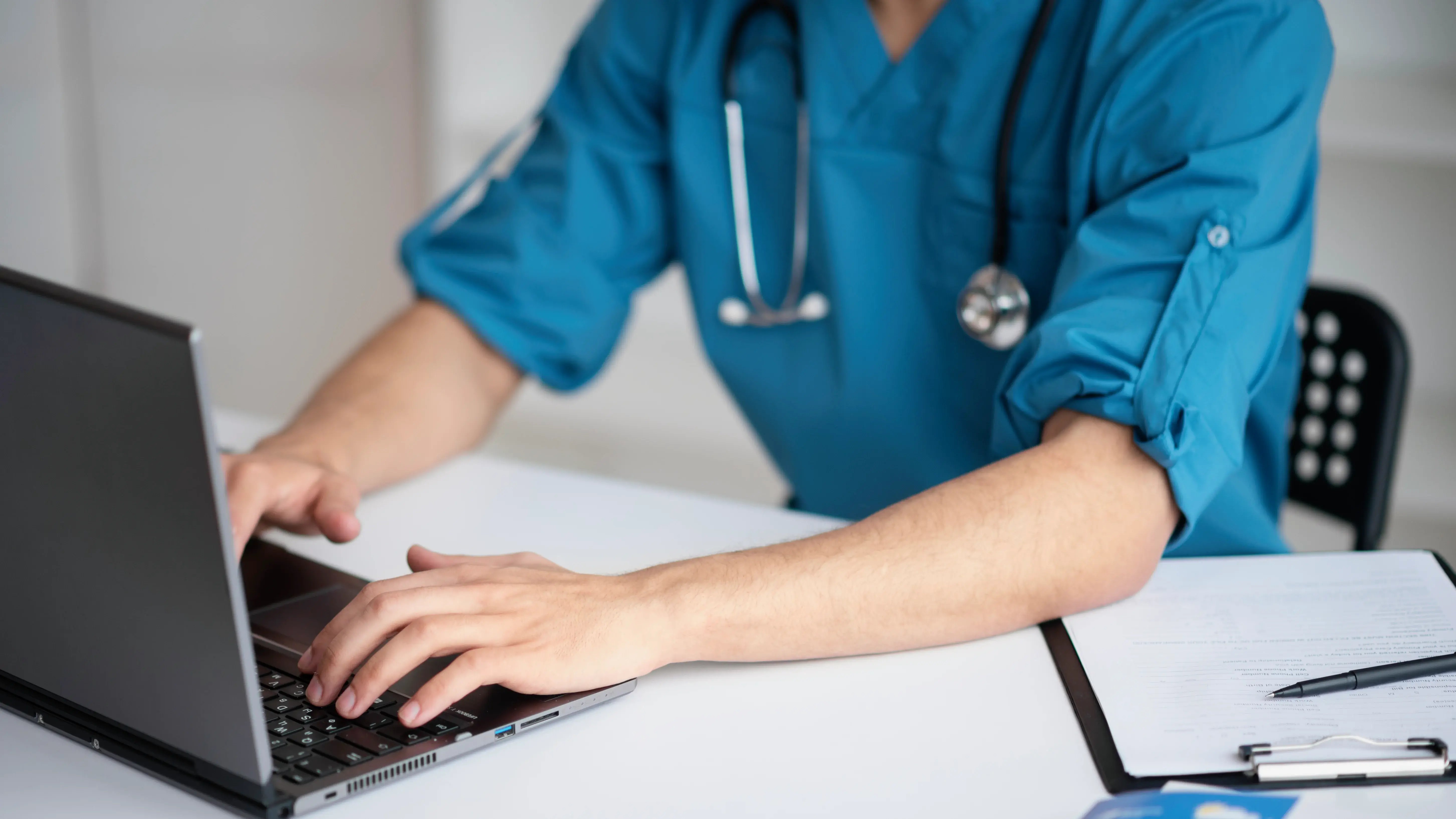 A doctor wearing a stethoscope, typing on their laptop while sitting